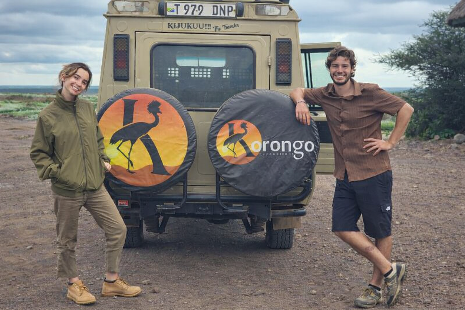 Two travellers standing beside a Korongo Expeditions safari vehicle, its spare wheel covers painted with the company's crowned crane logo